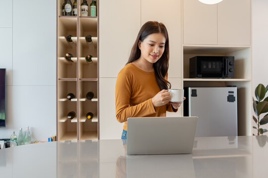Smiling Asian Woman Working on Laptop with Coffee in Kitchen