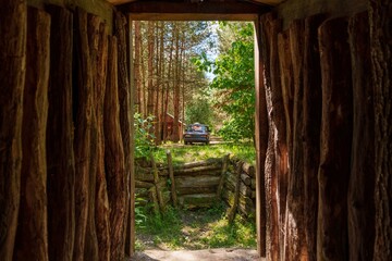 Fototapeta premium Rustic wooden log shelter interior with doorway view of forest pine trees sunlight and small wooden fence with parked car outside