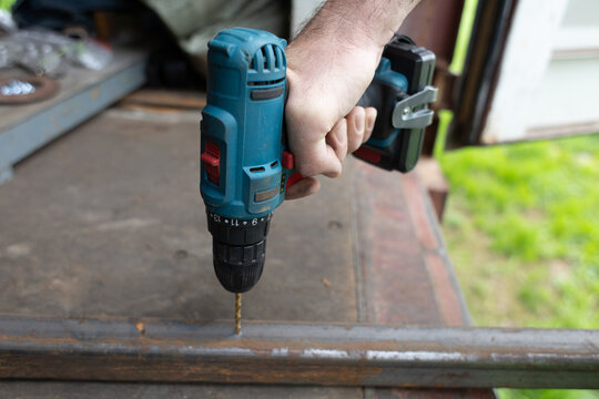 A close-up of a hand holding a cordless drill. The drill is positioned above a metal surface, ready to drill. The background is blurred with green grass visible.