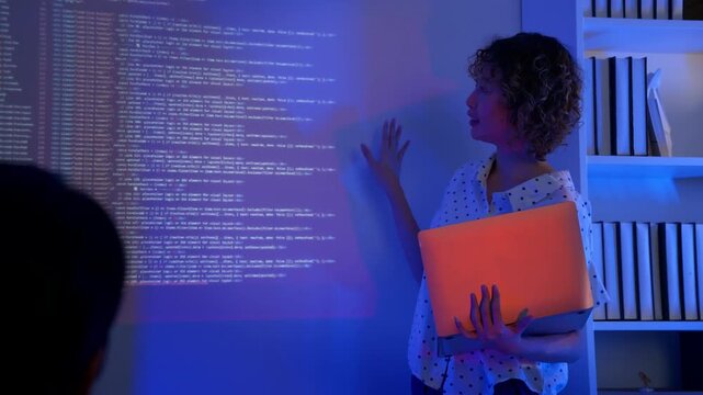 Young Asian female software developer holding laptop presents programming code on large projected screen with blue light during code review session. Concept of women in tech, modern tech mentorship.