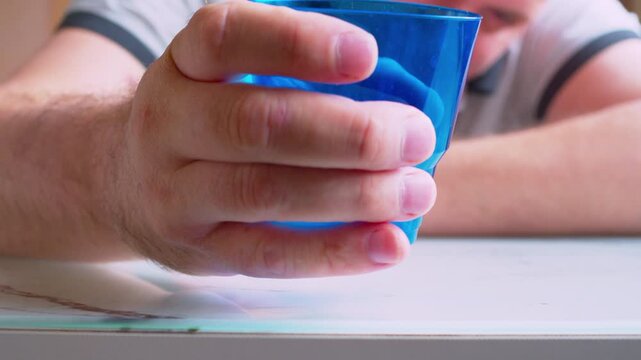 Close-up of a hand crushing an empty plastic cup, blurred silhouette of a drunk man sitting background. Cinematic slow-mo shot of an intoxicated man squeezing and destroying a disposable cup. 