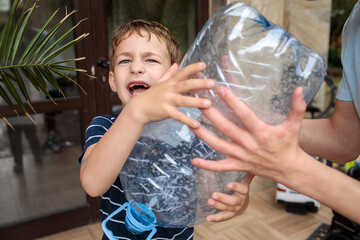 Obraz premium Little boy in navy striped shirt grabs a large plastic water bottle with both hands making a funny grimacing face near a palm plant with teen brother's hands visible beside him