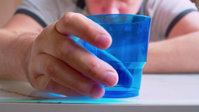 Hand holding empty plastic cup, blurred silhouette of drunk man sitting at a table. Close-up, shallow DoF, natural light. Metaphor for alcohol addiction, loneliness and despair.