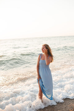 Young adult woman 30-35 year old wearing blue dress standing in water at beach over sea at background. Summer vacation time. 30s.