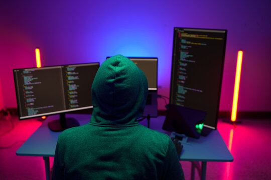 Hooded programmer working at multiple monitors in neon light
