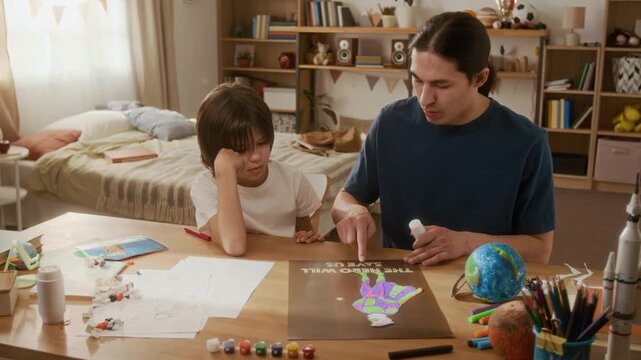 Asian young father helping multiethnic boy with school art project at home, gluing paper elements on poster. Footage ideal for parenting, family bonding, and creative education commercials