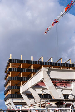 Crane arm crosses cloudy sky over timber structure, concrete frame and construction building, showing unfinished floors and urban worksite height