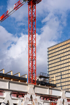 Red crane lifts above construction frame beside office building, linking timber structure, machinery and urban development under broken cloud