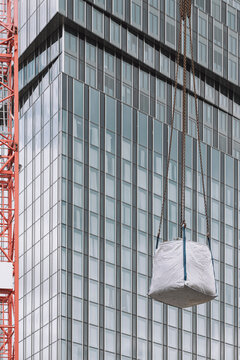 Hoisted load hangs from red crane beside glass tower facade, revealing construction lift, worksite scale and dense urban building detail