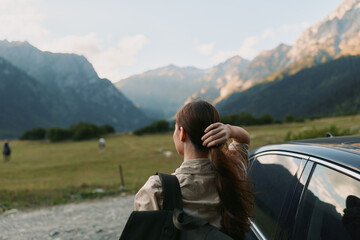 Fototapeta premium Young woman with long hair stands by a car in a mountain valley, looking at the distant peaks during a calm late afternoon.