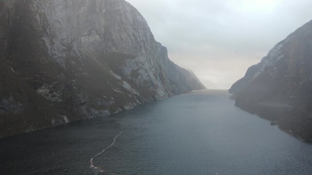 Dramatic 1080p drone footage flying through the misty and foggy Lysefjord canyon in Norway. High vertical mountain cliffs rise from the dark water under overcast skies. Real time 30fps.