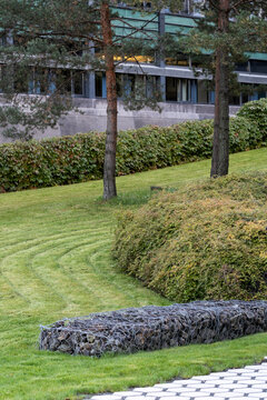 Landscaped lawn curves through urban garden with trees, hedges and stone edging, framing residential building grounds in soft daylight