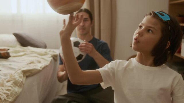 Asian man and young multiethnic boy spinning soccer ball and planet model on fingers while playing in bedroom. Footage is ideal for family lifestyle commercials, parenting campaigns