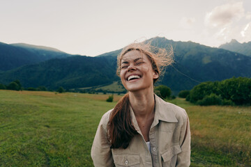 Obraz premium Cheerful woman outdoors, smiling with eyes closed, sunlit moment in a field near mountains, casual jacket, light breeze, natural colors, lush greenery, carefree mood today