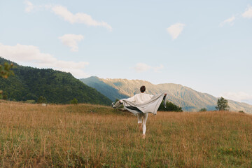 Fototapeta premium Young woman stands in a sunlit field with arms outstretched, a flowing shawl catching the breeze, while distant mountains frame a peaceful, expansive countryside scene.