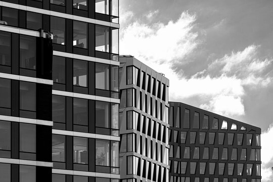Cloudy sky opens above office blocks in a black and white photograph, where patterned facade and windows layer urban architecture depth