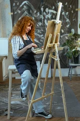 Fotobehang Schilderij female painter creating artwork with paintbrush on canvas standing near easel in authentic art studio interior. concept of creativity, inspiration and artistic profession  © Andrii Lysenko