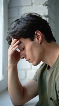 Young man in olive green t-shirt touching forehead with eyes closed near window close up