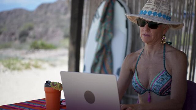 Mature woman works on computer at tropical beach while showing various emotions as she enjoys her vacation time