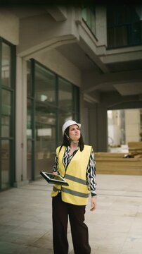 A woman engineer in a hard hat and safety vest surveys a construction site. She uses a digital tablet for inspection and project management