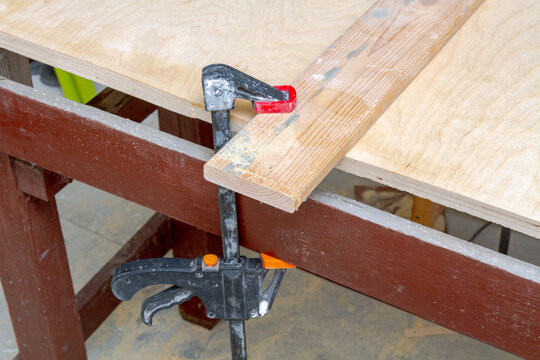 Wood plank secured with a metal bar clamp on a workbench in a workshop. Carpentry tool for holding timber pieces during assembly. Manual labor, home renovation and woodwork project task