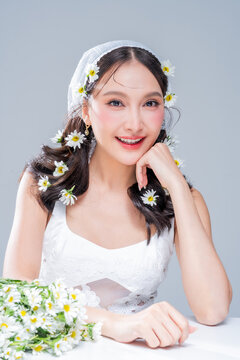 Charming Asian woman resting chin on hand with daisy flowers on table for mindful skincare cosmetics campaign slow living aesthetic fairycore romantic portrait wellness mindfulness studio