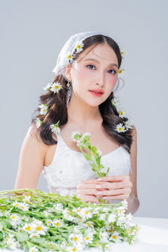 Charming Asian woman resting chin on hand with daisy flowers on table for mindful skincare cosmetics campaign slow living aesthetic fairycore romantic portrait wellness mindfulness studio