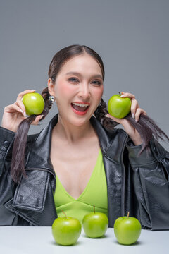 Excited Asian woman holding green apples by braids for dynamic nutrition diet marketing campaign neon street fashion aesthetic healthy lifestyle vibrant portrait studio
