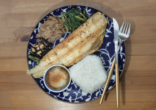 An elegantly composed Japanese set meal features a beautifully grilled, golden-brown Shima Hokke fillet resting diagonally across a deep-blue patterned ceramic plate. In the foreground, a clean, squar
