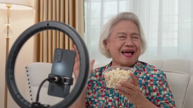 Elderly Woman's Delight: A joyful elderly woman beams with laughter, clutching a bowl of popcorn in her hand, framed by a soft ring light, as she engages with a smartphone.