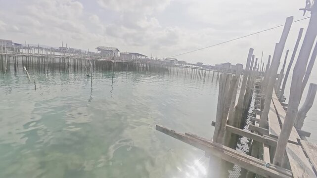Wooden pier extending across calm sea water toward a coastal stilt house settlement under bright daylight and dramatic cloud formations