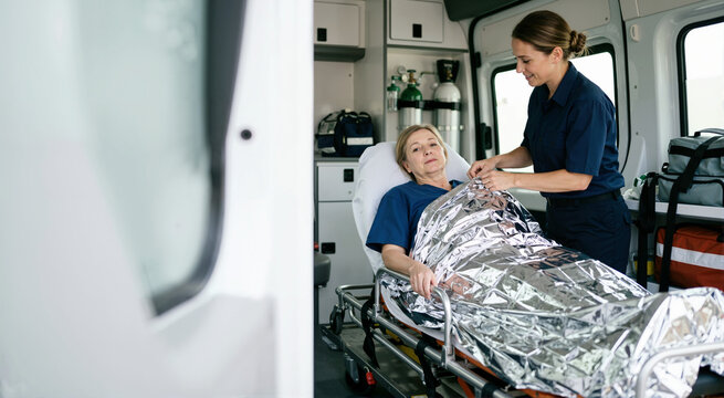 Female paramedic covering a patient with a silver thermal emergency blanket inside an ambulance. Emergency medical technician providing first aid and care during transport. Healthcare and rescue