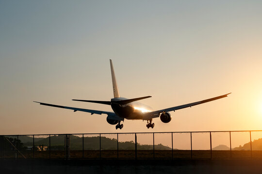 A wide-body airliner approaches an airport runway for landing. Its landing gear is deployed as it flies low over a security fence during a beautiful sunset
