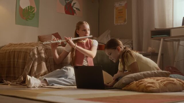 Stab shot of girls sitting on floor, older child practicing flute while younger using laptop during online music lesson. Footage ideal for promoting e-learning platforms and remote music tutoring