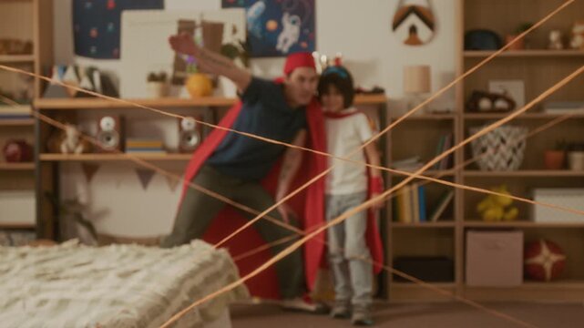 Rack focus shot of young man and child wearing superhero capes playing in bedroom with yarn laser maze. Ideal for promoting family bonding and creative parenting concepts