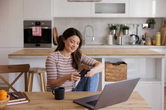 Woman Using Smartphone While Sitting at Kitchen Table Laptop is nearby