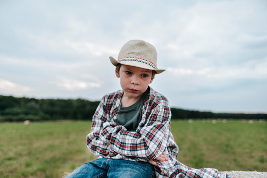 Young Boy in a Hat Sits Pensively on a Fence in a Green Field