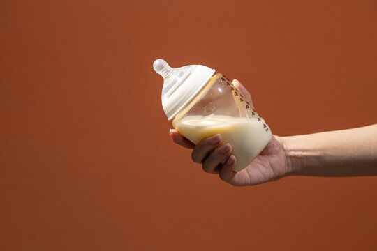 Hand holding a baby bottle filled with milk against a warm brown