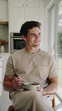 Young man in beige t-shirt eating bowl of granola in modern kitchen interior, medium shot