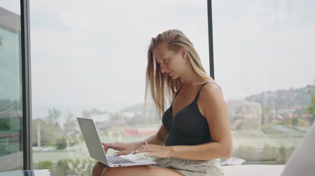 Young woman focuses on her laptop typing an email, working remotely from a sofa by a large window with stunning tropical landscape views