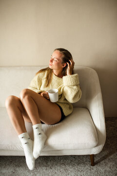 Young adult woman wearing knitted sweater and socks drinking coffee resting in couch at home in room. Relaxation. Cozy atmosphere.