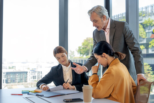 Experienced senior executive manager giving professional advice and guidance to female colleagues during a serious business document review in a modern office boardroom