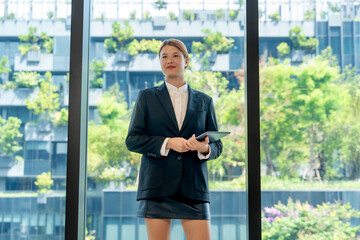 Fototapeta premium Portrait of a confident and successful young businesswoman in black suit holding a digital tablet near modern office window with visionary expression