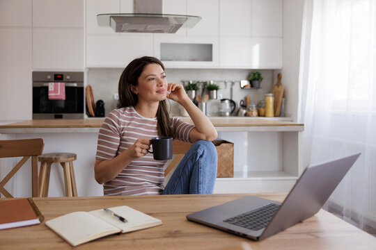 Woman Sits in Kitchen With Cup of Coffee and Laptop During Morning