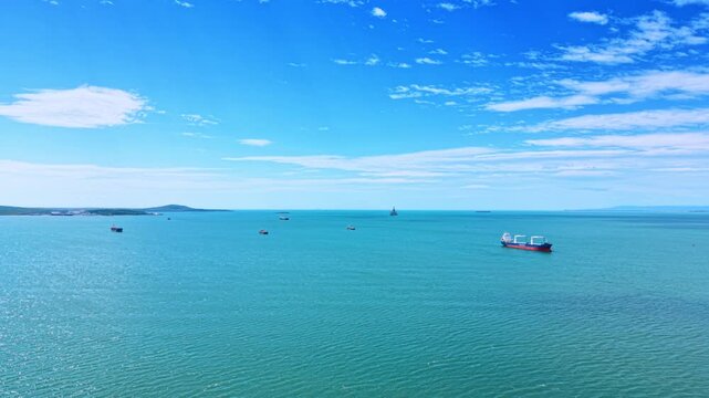 Aerial view of cargo ships anchored in turquoise sea. High angle shot of cargo vessels floating on calm turquoise water under a blue sky with white clouds.