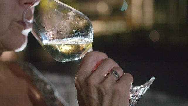Close-up of a senior woman holding and drinking crisp white wine from a glass at night. The backlight highlights her hand and the condensation on the cool beverage. She sips the drink