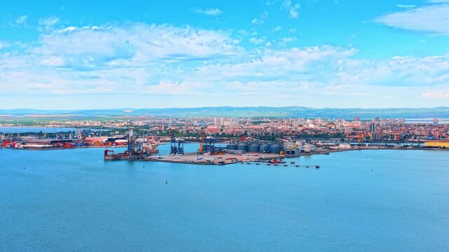 Industrial seaport infrastructure with container terminal and cargo cranes. Aerial view of a busy commercial port harbor with grain silos, shipping cranes, and city skyline.
