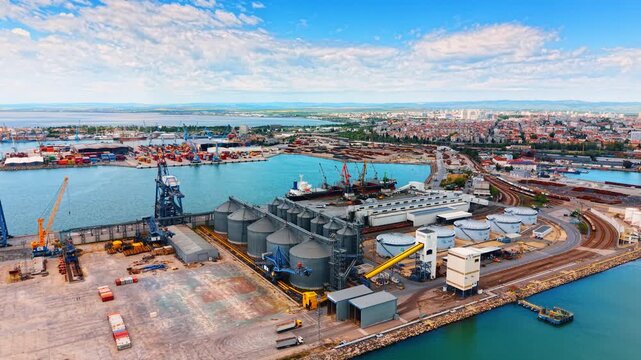 Waterfront grain silos. Workers move equipment near grain silos at a busy port. Ships are docked and containers are stacked on land.