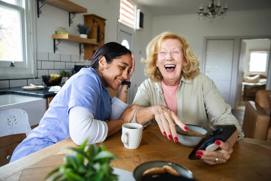 Caregiver and senior woman laughing over smartphone in home kitchen