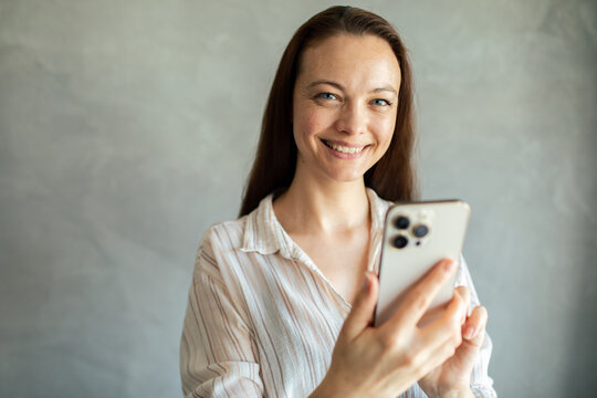 Smiling woman using smartphone indoors against gray wall